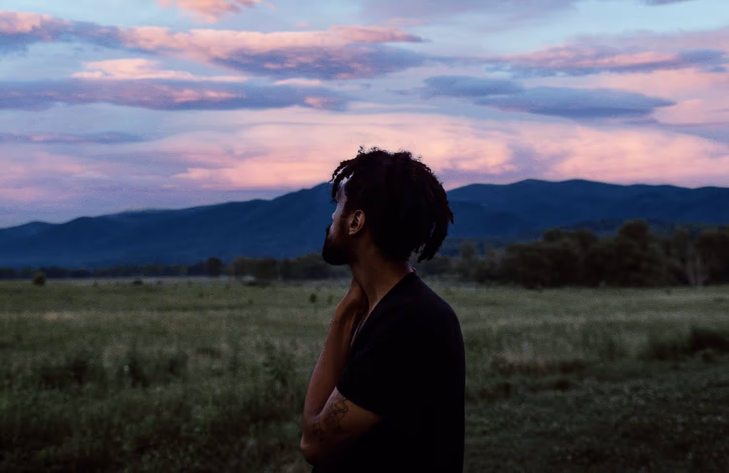 Film Photo of a man standing in a green field at dusk, looking at a sky of painterly filmic colors. Shot by Ivana Cajina